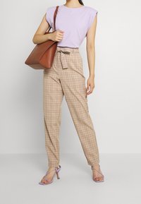 Kvinna i lila ärmlös topp, beige rutiga högmidjade byxor med bälte, lila klackar, bärande en stor brun läderhandväska på axeln.