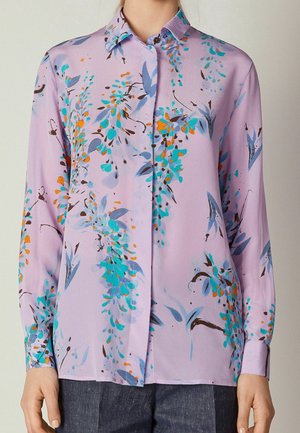 Lavendelfarbene Bluse mit langen Ärmeln und einem Kragen, mit einem Blumenmuster in Türkis, Orange und Dunkelbraun, getragen zu einer dunklen Hose.