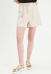 Shorts en coton beige avec une taille à cordon, des poches latérales et une coupe décontractée. Associés à des sandales dorées à lanières sur un fond blanc.