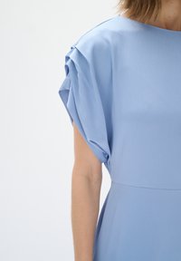 Abito azzurro chiaro in tessuto testurizzato, con maniche corte plissettate e una vestibilità rilassata, che mette in risalto un design liscio e minimalista.