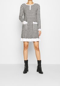 Vestito a scacchi bianco e nero con maniche lunghe, tasche e scollatura rotonda, con orlo arricciato e chiusura con bottoni.