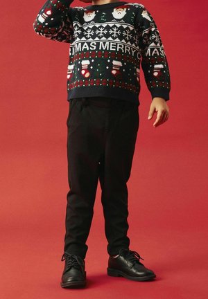 Enfant portant un pull de Noël festif avec le Père Noël et des bas de Noël, un pantalon noir et des chaussures noires sur un fond rouge.