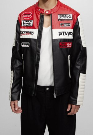Personne portant une veste de style course noire, rouge et blanche avec plusieurs écussons de sponsors et une chemise blanche en dessous.