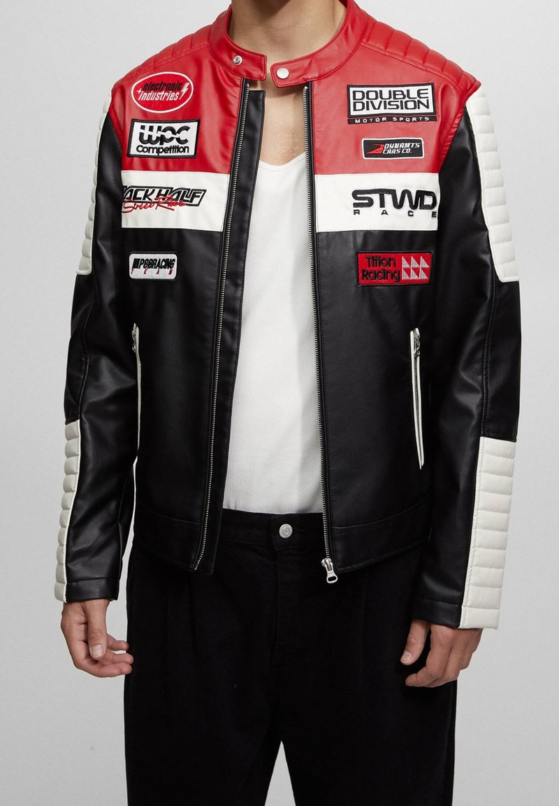Personne portant une veste de style course noire, rouge et blanche avec plusieurs écussons de sponsors et une chemise blanche en dessous.