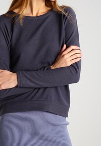 Kvinna i en mörk långärmad topp och en ljuslila ribbad kjol, med armarna korsade vid midjan framför en ljus bakgrund.