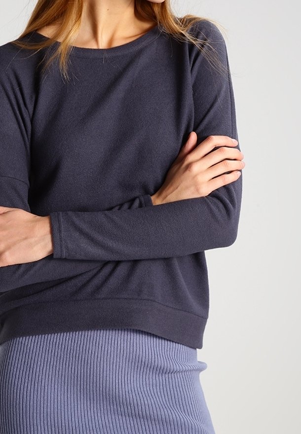 Kvinna i en mörk långärmad topp och en ljuslila ribbad kjol, med armarna korsade vid midjan framför en ljus bakgrund.