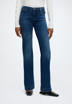 Femme portant un jean bleu foncé à jambes droites, un haut blanc à manches longues et des chaussures plates noires, debout sur un fond blanc.