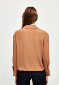 Lange mouw blouse in lichtbruin, gladde stof, met een kraag en knoopsluiting bij de manchetten, gedragen met donkerdenim broek.
