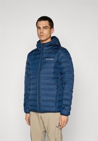 Mand iført en blå quiltet Columbia hættejakke og beige cargo bukser, står mod en ensfarvet lys grå baggrund.