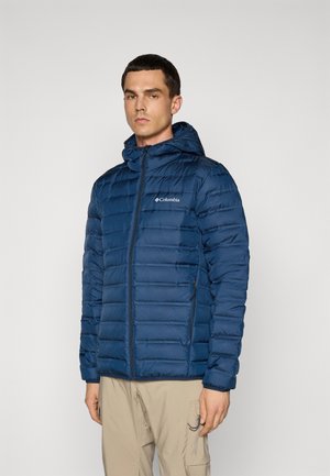 Man in een blauwe gewatteerde Columbia capuchonjack en beige cargobroek, staand tegen een effen lichtgrijze achtergrond.