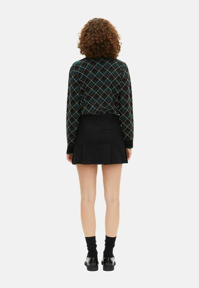 Žena s kučeravými vlasmi, oblečená v tmavom károvanom svetríku, čiernej plisovanej mini sukni, čiernych ponožkách a lesklých čiernych topánkach, otočená chrbtom na bielom pozadí.