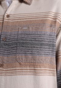 Camicia beige con righe orizzontali arancioni e grigie. Presenta una tasca sul petto con un'etichetta logo e chiusure a bottoni lungo la parte anteriore.