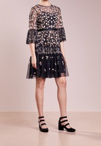Robe noire brodée de fleurs avec des manches transparentes et une jupe à volants ; assortie à des talons en daim noir avec plusieurs brides de cheville.