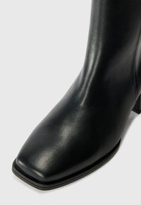 Botín de cuero negro hasta el tobillo con punta cuadrada, textura suave y un tacón modesto, que presenta un diseño elegante y minimalista.