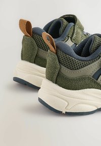 Zielone sportowe buty wyróżniają się siateczkowymi i zamszowymi materiałami, posiadają wyściełany kołnierz, brązową pętelkę z tkaniny oraz teksturowaną białą podeszwę środkową.