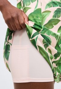 Ręka podnosząca zieloną tkaninę w liściasty wzór, odsłaniając pod spodem jasnoróżowy strój z kieszeniami na górnej części uda.