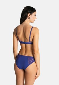 Femme portant un ensemble de lingerie bleu assorti, avec des détails en maille et en dentelle, se tenant de dos face à la caméra sur fond blanc.