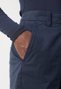 Mörkhyad hand som vilar i fickan på marinblå byxor som bärs med en matchande långärmad skjorta mot en ljus bakgrund.