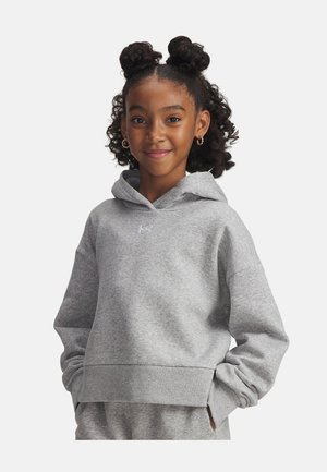 Smiling girl with curly hair in two buns wearing gold hoop earrings and a light gray hoodie with hands in pockets against white background.