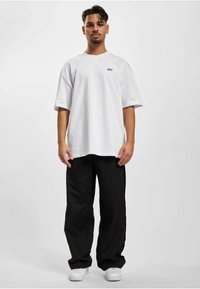 Hvid oversized t-shirt med grøn "DEF" logo, stylet med sorte vide bukser og hvide sneakers, præsenteret på en neutral baggrund.