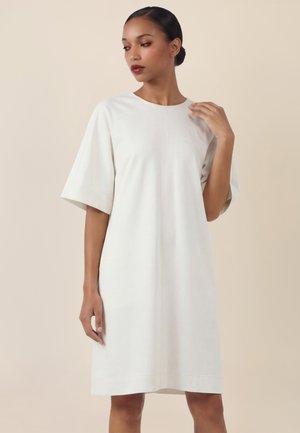 Kobieta z ciemnymi włosami związanymi w niski kok, ubrana w luźną, krótką sukienkę białą do kolan, patrząca lekko w dół i na bok.