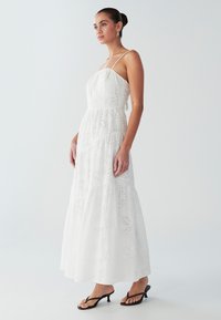 Robe longue blanche à volants, en coton texturé avec motifs ajourés, bretelles fines et corsage ajusté. Tongs noires portées.