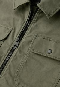 Oliwkowozielona kurtka z zamkiem błyskawicznym, dwoma przednimi kieszeniami i kołnierzem; wykonana z teksturowanej tkaniny z czarnymi elementami ozdobnymi.