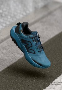 Plavajno-crne New Balance tenisice za trčanje s Gore-Tex tkaninom u zraku iznad asfaltirane ceste, blizu rubnika i taktilne staze.