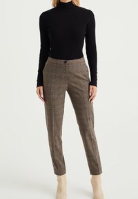 Black ribbed turtleneck paired with brown plaid tailored trousers. Trousers feature a button closure and tapered legs, worn with beige ankle boots.