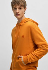 Oranje hoodie van zachte stof, met een voorzak, rib gebreide mouwen en een klein vierkant logo op de borst.