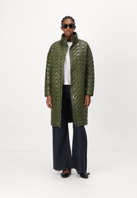 Grüner, gesteppter langer Mantel mit hohem Kragen und wellenförmiger Textur. Kombiniert mit weit geschnittenen, dunklen Jeans und schwarzen Slippern.