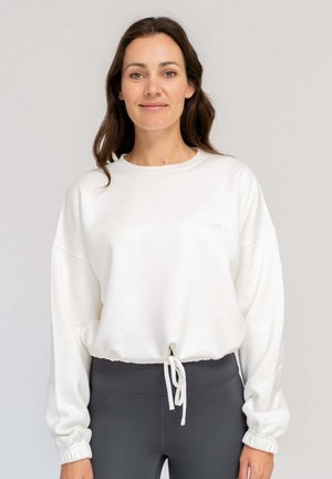 Lächelnde Frau mit langen braunen Haaren, die einen weißen Sweatshirt mit Kordelzug und dunkle, hoch taillen Leggings vor einfachem Hintergrund trägt.