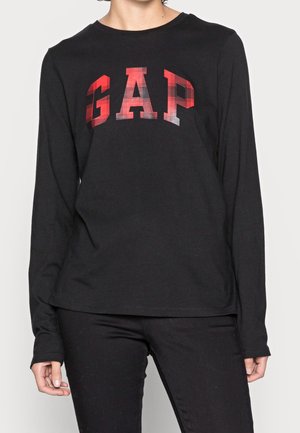 Personne portant un haut noir à manches longues avec des lettres à carreaux rouges "GAP" sur la poitrine et un pantalon noir sur fond uni.