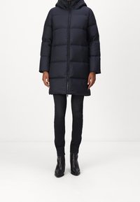 Musta toppatakki, jossa huppu, vetoketjukiinnitys edessä ja vaakasuora tikattu muotoilu. Yhdistetty tummiin kapealahkeisiin farkkuihin ja mustiin nilkkureihin.