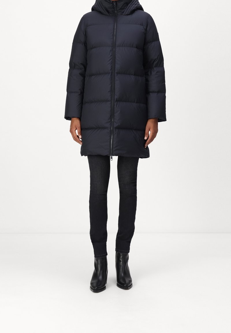 Musta toppatakki, jossa huppu, vetoketjukiinnitys edessä ja vaakasuora tikattu muotoilu. Yhdistetty tummiin kapealahkeisiin farkkuihin ja mustiin nilkkureihin.