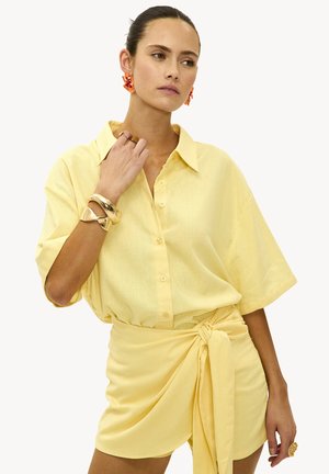 Femme avec les cheveux plaqués en arrière portant une chemise jaune à boutons et manches courtes, ainsi que des shorts noués assortis, accessoirisée avec des bijoux en or et des boucles d'oreilles orange.