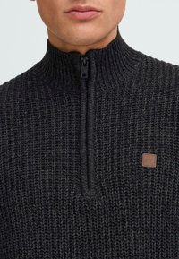 Dunkelgrauer Rippenstrickpullover mit halbem Reißverschluss, strukturiertem Finish und kleinem braunen Logo-Patch auf der Brust.