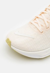 Chaussure de course Nike beige clair avec logo swoosh blanc, tige en maille et lacets, montrant les détails avant et latéraux.