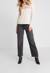 Maglione beige a maniche lunghe con motivi geometrici rossi, abbinato a jeans neri a gamba larga e stivali neri alla caviglia con punta affusolata.