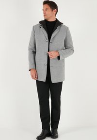 Šedý vlněný kabát s černou kapucí, se zapínáním na knoflíky, s vykrojeným límcem a postranními kapsami, kombinovaný s černými kalhotami a botami.