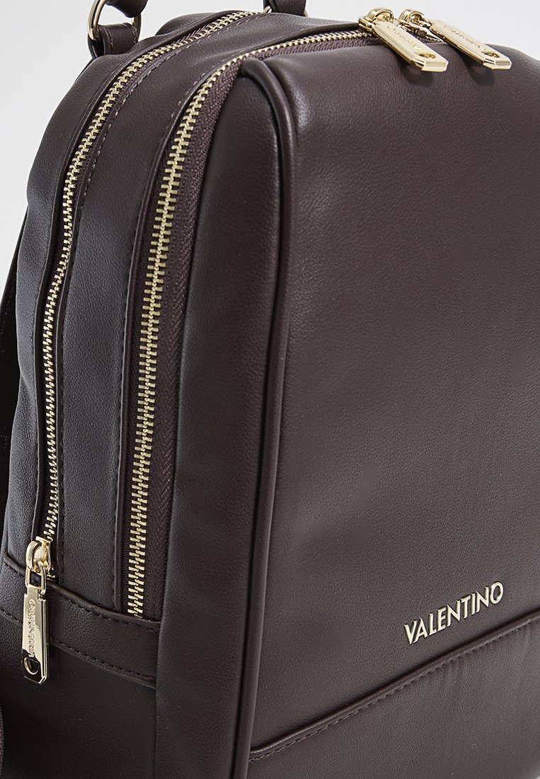 Brauner Leder-Rucksack mit goldfarbenen Reißverschlüssen und Beschlägen. Glatte Textur, leicht strukturiertes Design, das den Markennamen in Gold präsentiert.