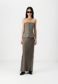 Top bez ramiączek w oliwkowym kolorze z peplum i zapięciem na guziki, w zestawieniu z dopasowaną, długą spódnicą. Modelka nosi czarne okulary przeciwsłoneczne i błyszczące czarne buty.