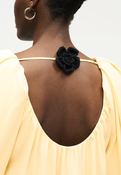 Η γυναίκα από πίσω, φοράει ανοιχτό κίτρινο φόρεμα με βαθύ στρογγυλό ντεκολτέ, λεπτό ιμάντα στους ώμους διακοσμημένο με ένα μαύρο πλεκτό λουλούδι.