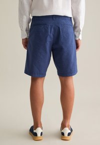 Pantalones cortos texturizados en azul marino para hombre, de longitud a la rodilla, con dos bolsillos traseros. Combinados con deportivas en azul marino con detalles en blanco y suela de goma.