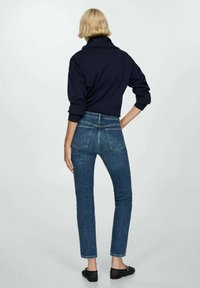 Pull bleu foncé avec un col montant et des manches longues, associé à un jean denim foncé ajusté, avec des détails de poches et une coupe droite.