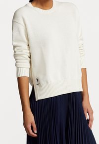 Suéter de punto blanco con cuello redondo y dobladillo acanalado. Presenta mangas largas y un pequeño logo en azul marino en la esquina inferior izquierda.