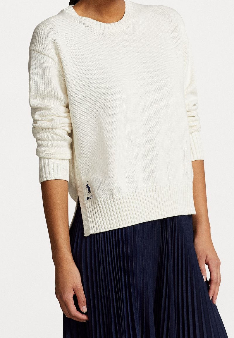 Suéter de punto blanco con cuello redondo y dobladillo acanalado. Presenta mangas largas y un pequeño logo en azul marino en la esquina inferior izquierda.