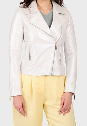Femme portant une veste en cuir blanche de motard avec des fermetures éclair argentées sur un haut gris, associée à un pantalon taille haute jaune pâle.
