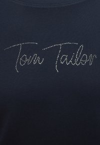 Ύφασμα ναυτικού μπλε με την επιγραφή «Tom Tailor» γραμμένη με καλλιγραφικά γράμματα χρησιμοποιώντας μικρές ασημένιες τρουκς ή στρας.
