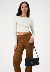 Ilgi piedurkņu krēmkrāsas topiņš, kas saskaņots ar brūnām ribotām plaša kāju biksēm. Modelis tur nelielu melnu rokassomu ar zelta sprādzi.